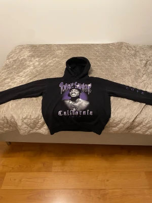 Svart Tupac Shakur hoodie California Love - Svart hoodie med tryck av Tupac Shakur och texten 'California Love' i lila och vitt. Hoodien har huva och långa ärmar med tryck även på ena ärmen. Perfekt för dig som gillar streetwear och hiphopstil. Även en hoodie för att ha hemma och ba chilla, ällmänt en bra hoodie och fin.