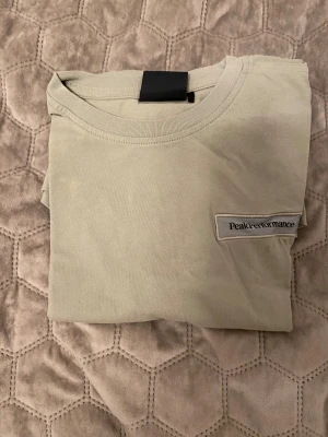 Beige Peak Performance t-shirt - Snygg beige t-shirt från Peak Performance med diskret logotyp på bröstet. Klassisk rund hals och korta ärmar. Tillverkad i mjuk bomull som känns skön mot huden. Perfekt för dig som gillar stilrena och enkla plagg.