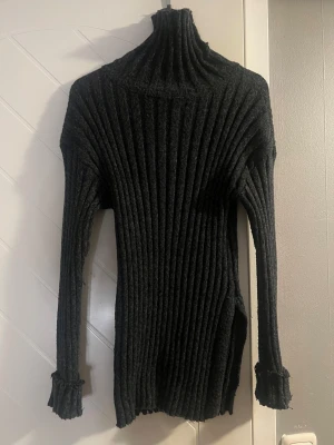 Mörkgrå ribbad polotröja med lång ärm - Säljer en mörkgrå ribbad polotröja med högt krage och långa ärmar. Tröjan är stickad i ett mjukt material och har en slim passform som sitter snyggt mot kroppen. 