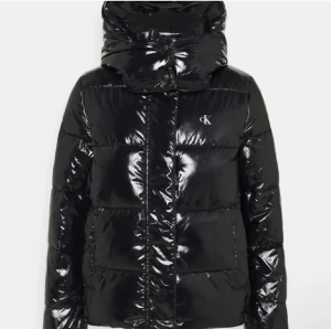 Svart pufferjacka från Calvin Klein - Säljer en svart, glansig pufferjacka från Calvin Klein med stor huva och CK-logga på bröstet. Jackan har quiltad design, dragkedja och knappar framtill. Perfekt för kalla dagar och har en riktigt modern vibe. Passar dig som gillar streetstyle och vill sticka ut.