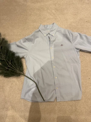 Ljusblå skjorta från GANT - Snygg ljusblå skjorta från GANT med klassisk button-down krage och broderad logga på bröstet. Skjortan har långa ärmar, vita knappar och är tillverkad i mjuk bomull. Perfekt nu när jul och nyår är runt hörnet