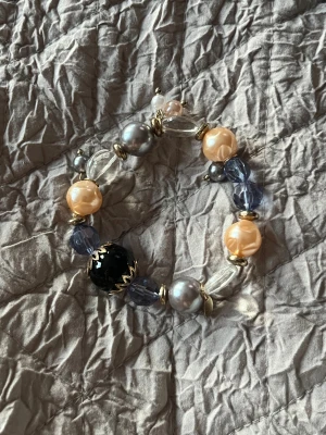 Färgglatt pärlarmband med mixade pärlor - Unikt armband med stora pärlor i silver, guld, blått, svart och beige. Armbandet har både glansiga och transparenta pärlor i olika former och storlekar, samt guldfärgade detaljer som ger en lyxig känsla. Perfekt för dig som vill sticka ut med accessoarerna!