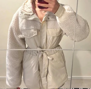 Beige teddyjacka med bälte - Säljer en supermysig beige teddyjacka med fluffiga ärmar och detaljer. Jackan har knappar framtill, två bröstfickor och ett knytbart bälte i midjan för snygg passform. Perfekt för kalla dagar och ger en trendig look.