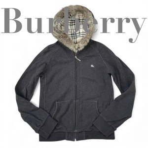 Burberry Black Label hoodie med päls - Mörkgrå hoodie från Burberry Black Label med dragkedja, ikonisk rutig huva fodrad med mjuk pälsdetalj. Jackan har två fickor fram och ribbade muddar. Materialet är bomull och huvan har äkta päls. Perfekt för dig som gillar klassisk stil med lyxig touch.