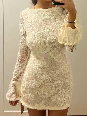 Vit spetsklänning med öppen rygg - Aldrig använd superfin kort vit spetsklänning med blommiga 3D-detaljer, långärmad med utsvängda ärmslut och en snygg öppen rygg. Klänningen har rund halsringning och dragkedja bak.