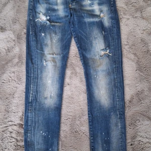 Blå jeans från Dsquared2 stl 44 - Snygga blå jeans från Dsquared2, modell 2112, med coola slitningar och blekta detaljer. Jeansen har klassisk femficksdesign, röd Dsquared2-tag vid fickan och är tillverkade i bomull med lite lycra för extra stretch. Perfekt för dig som gillar en edgy streetstil.