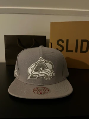 Grå keps Mitchell & Ness  - Snygg grå keps från Mitchell & Ness med Colorado Avalanche-logga broderad framtill i vitt och silver. Klassisk flatbrim och justerbar passform. Perfekt för dig som gillar sportiga vibes och vill ha en clean look.
