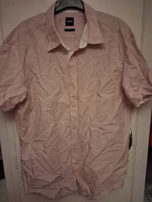 Ljusrosa skjorta från Hugo Boss  - Snygg ljusrosa skjorta från Hugo Boss i storlek XL. Skjortan har klassisk krage, korta ärmar och knäppning framtill med vita knappar. Materialet är mjuk bomull med diskret struktur i tyget som ger en cool touch. Perfekt för dig som gillar stilrena och fräscha plagg.