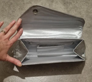 Silvrig kuvertväska i krokopräglat skinn - Snygg kuvertväska i metallic-silver med krokodilpräglat mönster. Väskan har ett stilrent snäpplås och insida i matchande silverfärgat tyg. Perfekt för dig som vill sticka ut med en trendig accessoar.