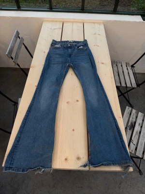 Blå bootcut jeans från 157 - Ett par klassiska blå jeans från 157 med bootcut-modell och slitna, fransiga benslut. Jeansen har fem fickor, hög midja och är tillverkade i ett mjukt denimtyg. Perfekta för dig som gillar en retro vibe och vill ha ett par jeans med lite extra attityd.