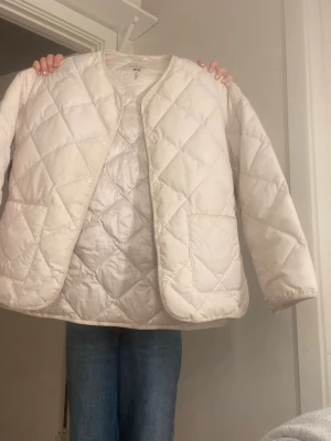 Vit pufferjacka Uniqlo - Säljer en vit pufferjacka från Uniqlo med quiltat mönster och rundad hals. Jackan har tryckknappar framtill, två fickor och är tillverkad i vattenavvisande syntetmaterial. Perfekt för dig som gillar clean och enkel stil.