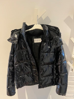Svart pufferjacka från Calvin Klein Jeans - Snygg svart pufferjacka från Calvin Klein Jeans med glansig finish och huva. Jackan har quiltad design, dragkedja framtill och logga på bröstet och ärmen. Perfekt för kalla dagar när du vill ha både stil och värme.