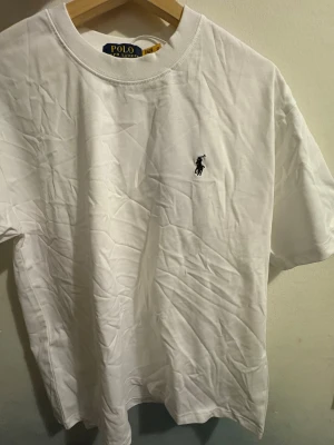 Vit t-shirt från Polo Ralph Lauren - Klassisk vit t-shirt från Polo Ralph Lauren med broderad svart logga på bröstet. T-shirten har rund halsringning och är tillverkad i mjuk bomull. Enkel och stilren design som passar perfekt till jeans eller shorts.