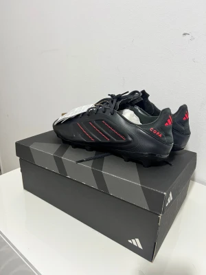 Adidas Copa Pure League fotbollsskor - Svarta Adidas Copa Pure League fotbollsskor med röda detaljer och klassiska tre ränder på sidan. Skorna har snörning och är tillverkade i skinn för optimal komfort och känsla på planen. Snygg, strömlinjeformad design med låg profil och platt sula med dobbar.