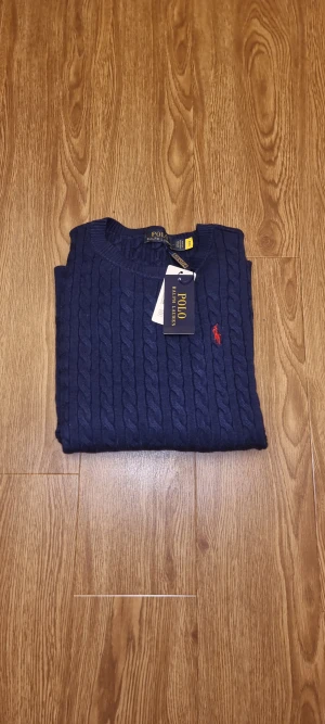 Marinblå stickad tröja Polo Ralph Lauren - Snygg marinblå stickad tröja från Polo Ralph Lauren med klassiskt kabelstickat mönster och rund hals. Tröjan har långärmad passform och den ikoniska röda logon broderad på bröstet. Perfekt för dig som gillar stilrena och tidlösa plagg.