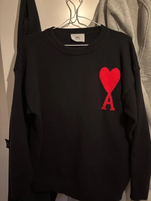 Svart stickad tröja från Ami - Svart stickad tröja från Ami Alexandre Mattiussi med röd hjärt- och A-logga på bröstet. Rund hals och ribbade muddar vid ärmslut och nederkant. Tillverkad i Portugal, perfekt för dig som gillar stilrena detaljer med en twist.