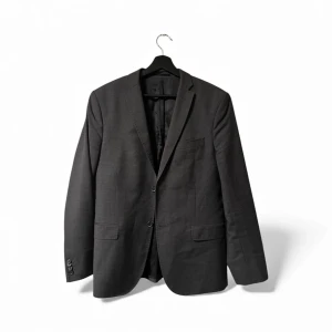 Mörkgrå kavaj från J.Lindeberg - Snygg mörkgrå kavaj från J.Lindeberg i slim fit-modell. Klassisk design med två knappar framtill, fickor med lock och slät yta. Fodrad insida i blankt material. Perfekt för dig som gillar stilrena och moderna plagg.