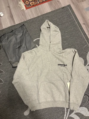 Grå Essentials Fear of God hoodie - Snygg grå hoodie från Essentials Fear of God med svart logotyp på bröstet och ryggen. Tröjan har huva, känguruficka och ribbade muddar. Mjuk insida och klassisk loose passform, perfekt för chill dagar. Kommer med originalpåse och tag.