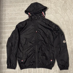 Svart vindjacka från Moncler med huva - Svart vindjacka från Moncler med huva och dragkedja framtill. Jackan har elastiska muddar, två fickor med dragkedja och Moncler-logga på ärmen. Snygga röda och vita detaljer vid huvan och fickorna. Perfekt för dig som gillar stilren och sportig look. Obs väldigt tunn
