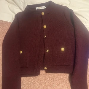 Vinröd kofta från Zara med guldknappar - Snygg vinröd kofta från Zara med guldfärgade knappar framtill och två fickor. Koftan är stickad och har en croppad passform med långa ärmar. Perfekt för dig som gillar klassiska detaljer och vill ha en stilren look.
