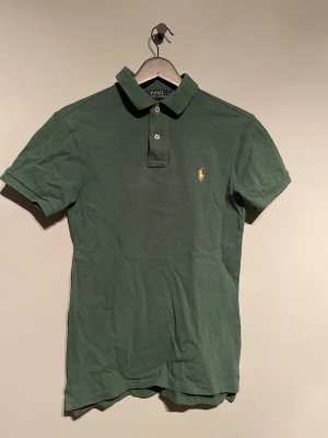 Grön pikétröja från Polo Ralph Lauren - Snygg grön pikétröja från Polo Ralph Lauren med klassisk krage och två knappar framtill. Tröjan har korta ärmar och den ikoniska gula broderade loggan på bröstet. Materialet är mjuk bomull som känns skönt mot huden. Perfekt för dig som gillar stilrena och tidlösa plagg.