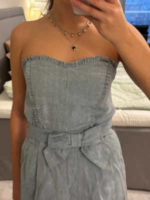 Sandro klänning - Ljusblå axelbandslös klänning i jeansliknande bomullstyg från Sandro. Klänningen har volangkant upptill, markerad midja med bredt knytband och stor rosett framtill. 