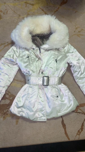 Vit pufferjacka med päls från Zara Woman - Säljer en glansig vit pufferjacka från Zara Woman med stor fluffig fuskpäls runt kragen. Jackan har bälte i midjan, dragkedja och knappar framtill. Perfekt för dig som vill ha en snygg och varm look under vintern. Den är i bra skick men behöver bara tvättas. (Använd) pris kan diskuteras 