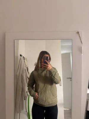 Olivgrön hoodie med ficka - Säljer en olivgrön hoodie med magficka och snörning i huvan. Tröjan har en avslappnad passform och är perfekt för dig som gillar en enkel och stilren look. Mjuk och skön, passar till jeans eller leggings.