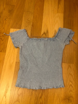 Ljusblå smockad topp från Stradivarius - Supersöt ljusblå topp från Stradivarius i smockad bomull med korta ärmar och offshoulder-design. Toppen har små volangdetaljer vid halsringning och nederkant samt knytband på ärmarna. Den har uttöjbart material. Perfekt för dig som gillar en feminin och trendig stil.