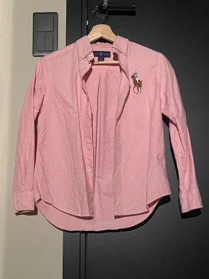Rosa skjorta från Ralph Lauren - Snygg rosa skjorta från Ralph Lauren i storlek 10-12 år. Klassisk design med knappar framtill, button-down krage och broderad logga med polospelare på bröstet. Tillverkad i mjuk bomull och har långa ärmar med manschettknappar. Perfekt för dig som gillar stilrena och tidlösa plagg.