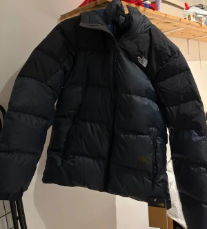 Svart pufferjacka från The North Face - Säljer en svart pufferjacka från The North Face med huva och dragkedja framtill. Jackan har ett stilrent, quiltat mönster och två sidofickor med dragkedja. Perfekt för kalla dagar och har en cool, oversized vibe. Har dock en fläck vid fickan men märks nästan inte. (Köpt på Tradera)
