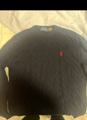 Mörkblå stickad tröja Polo Ralph Lauren - Säljer en mörkblå stickad tröja från Polo Ralph Lauren i storlek M. Tröjan har klassiskt kabelstickat mönster och en röd broderad logga på bröstet. Rund hals och långa ärmar, perfekt för dig som gillar stilren och tidlös design. Pris hugget i sten!