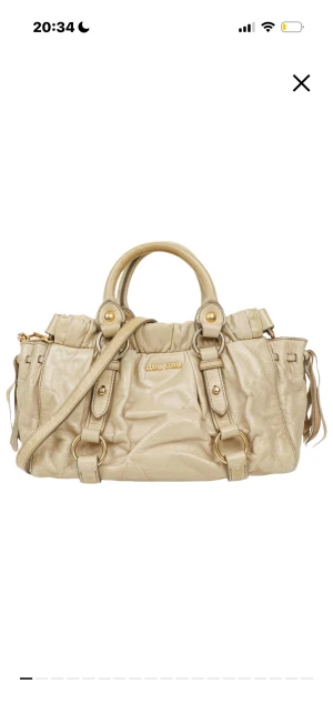 Beige Miu Miu väska - Snygg beige handväska från Miu Miu i mjukt skinn med guldfärgade metalldetaljer och dubbla handtag. Väskan har justerbar axelrem, dekorativa spännen och ringar samt rynkade detaljer upptill. Perfekt storlek för allt du behöver och riktigt lyxig vibe. Måtten på väskan är höjd- 16cm bredd-26cm djup-11,5cm. Väskan är köp på Vestiaire Collective och är äkta. Väskan har blivit kollad av professionella som garanterat äktaheten. Kvitto finns samt äktahetsbevis. Priset går att diskutera.🩷