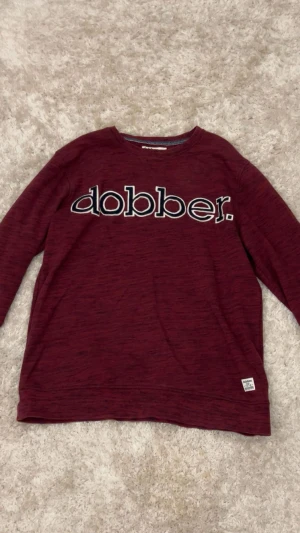 Vinröd långärmad tröja från Dobber - Snygg vinröd långärmad tröja från Dobber med stort broderat logotyptryck över bröstet. Rund halsringning och mjukt material som känns skönt mot huden. Perfekt för dig som gillar enkel och stilren design. Perfekt nu till vintern