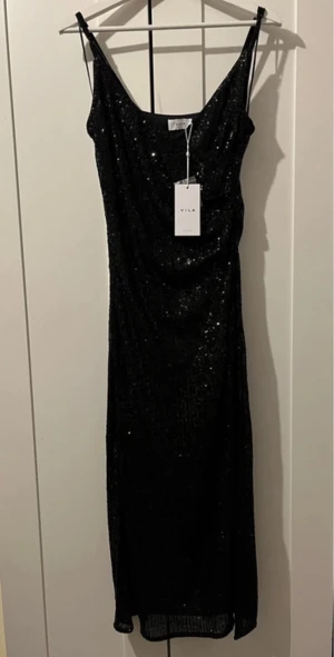 Svart glittrig långklänning från Vila - Svart långklänning från Vila med glittriga paljetter över hela klänningen. Klänningen har v-ringad hals, smala axelband och en snygg draperad passform. Perfekt för dig som vill sticka ut med en elegant och trendig look.