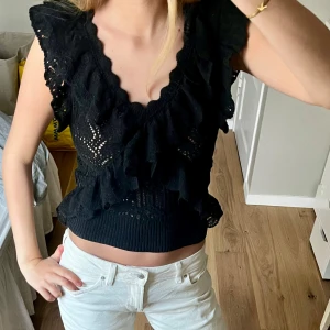 Svart volang topp - Snygg svart topp med volangdetaljer och v-ringning. Den är ärmlös och croppad, perfekt för en trendig look. Passar bra till jeans eller kjol.