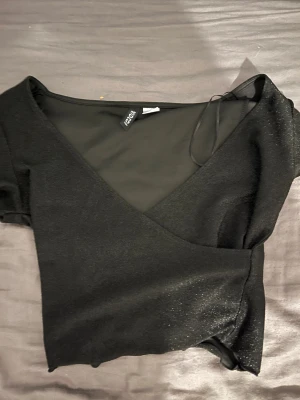 Svart omlottopp med glitter från HM - Svart omlottopp från HM med glittrig finish och djup v-ringning. Toppen har korta ärmar och knyt i sidan för en snygg passform. Perfekt för dig som gillar stilrena plagg med lite extra shine.