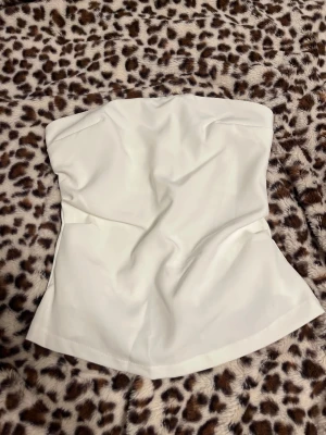 Vit tubtopp med dragkedja - Säljer en stilren vit tubtopp med axelbandslös design och lätt utsvängd form. Toppen har en diskret dragkedja baktill för enkel på- och avtagning. Materialet känns slätt och har en snygg struktur som ger en clean look. Perfekt för att skapa en chic och modern outfit.