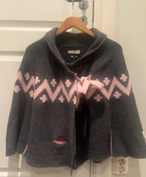 Mörkgrå poncho Odd Molly - Säljer en mörkgrå poncho från Odd Molly med ljusrosa zigzag-mönster och dekorativ rosett framtill. Jackan är stickad, har lång ärm och en mjuk krage. Perfekt för dig som gillar snygga detaljer och skön passform.