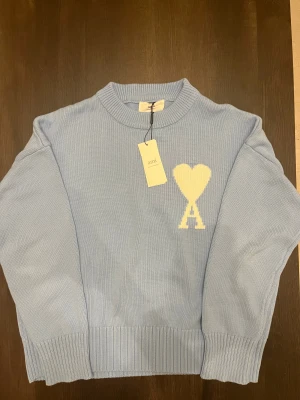 Ljusblå stickad tröja från Ami Paris - Snygg ljusblå stickad tröja från Ami med ett stort vitt hjärta och A på bröstet. Tröjan har rund hals, ribbade muddar och är långärmad. Perfekt för dig som gillar stilrena detaljer och vill ha något som sticker ut lite extra.
