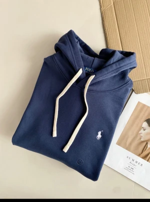 Mörkblå hoodie från Polo Ralph Lauren - Snygg mörkblå hoodie från Polo Ralph Lauren med vit broderad logga på bröstet och vita snören i huvan. Klassisk och stilren modell med mjukt material och avslappnad passform. Perfekt för dig som gillar enkel men ikonisk streetstyle.