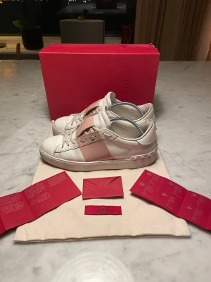 Valentino Garavani Open vita/rosa - Säljer ett par Valentino Garavani Open sneakers i vitt skinn med bred rosa panel över sidan. Snygga nitar längs hälen. Storleken är 40. Skorna har äktahetsbevis (se på bild 4) dustbag, box och OG medföljer. Skorna koster runt 7000 nypris. Skorna är använda ett fåtal gånger.                             Skriv vid frågor, priset är inte hugget i sten 🤝