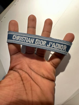 Blå Christian Dior J'Adior armband - Snyggt blått armband från Christian Dior med texten 'J'Adior' broderad i vitt. Armbandet har en cool, vävd design och känns riktigt lyxigt och trendigt. Perfekt accessoar för att lyfta din outfit med lite designer-vibe.