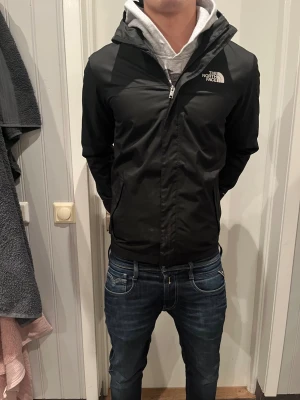  The North Face  - Säljer en svart vindjacka från The North Face med huva och vit logga på bröstet och ryggen. Jackan har dragkedja framtill, två sidofickor och är långärmad. Perfekt för dig som vill ha en stilren och funktionell jacka med snygg passform.
