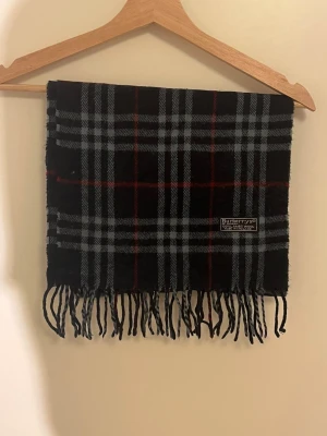  Halsduk från Burberry - Snygg halsduk från Burberrys i klassiskt rutig design med svarta, gråa och röda toner. Gjord av 100% lammull och har tvinnade fransar i ändarna. Perfekt för att ge din outfit en cool London-vibe och hålla dig varm under kyliga dagar. Den är såklart äkta och i väldigt bra skick! Skriv vid minsta fundering!