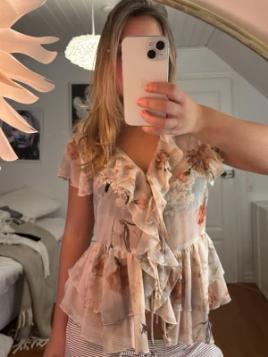 Blommig volangblus från Zara - Supersöt blommig blus från Zara i ljusa toner med volanger och knytband framtill. Blusen har korta ärmar, v-ringning och flera lager volanger som ger en romantisk vibe. Perfekt för dig som gillar en feminin och luftig stil.