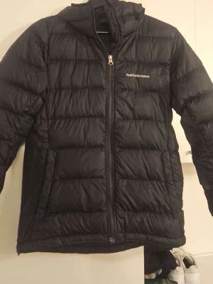 Svart dunjacka Peak Performance - Svart dunjacka från Peak Performance med huva och dragkedja framtill. Jackan har quiltad design och är långärmad, perfekt för kalla dagar. Snygg och enkel stil med logga på bröstet. Passar dig som gillar funktion och klassisk look. Sparsamt använd, Strolek S
