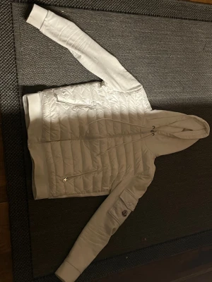 Vit pufferjacka från Moncler med huva - Säljer en snygg vit pufferjacka från Moncler i storlek M. Jackan har huva med snörning, quiltad framsida, dragkedjor och en ficka med Moncler-logga på ärmen. Materialet är polyester och jackan har en clean, modern look.