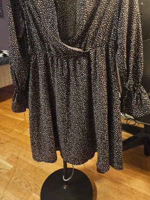 Svart prickig omlottklänning Shein - Svart kort omlottklänning från Shein med vita prickar, v-ringad hals och långa ärmar med volang vid ärmslutet. Klänningen har en luftig passform och är tillverkad i ett lätt syntetmaterial som känns skönt mot huden.