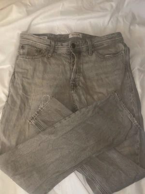 Grå relaxed jeans från Jack & Jones - Säljer ett par grå jeans från Jack & Jones, modell Relaxed/Chris. De har en loose passform med raka ben och klassisk femficksdesign. Jeansen är tillverkade i bomull. Dem har dock ett hål vid vänster bakficka (kolla bilden) men som borde gå att åtgärda!